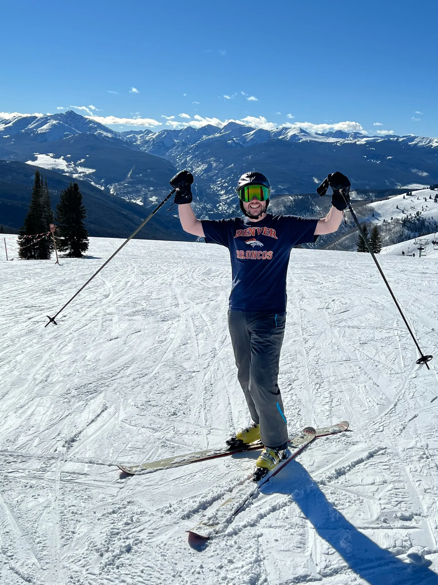 Parker on skis at the top of a run in Vail, poles raised, on a clear day with snowy peaks behind him.