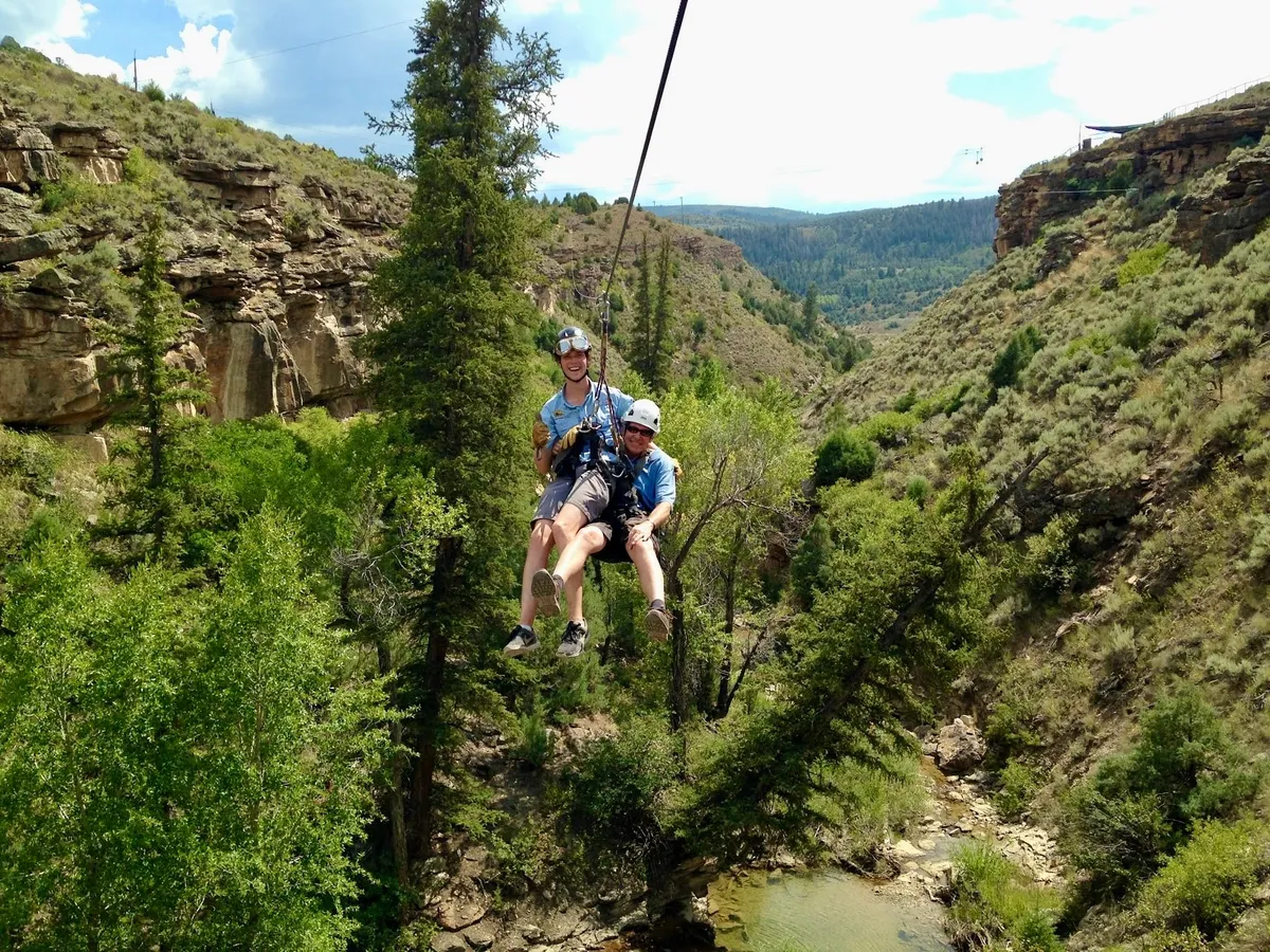 Parker on a zipline above a wooded canyon, alongside a second rider.